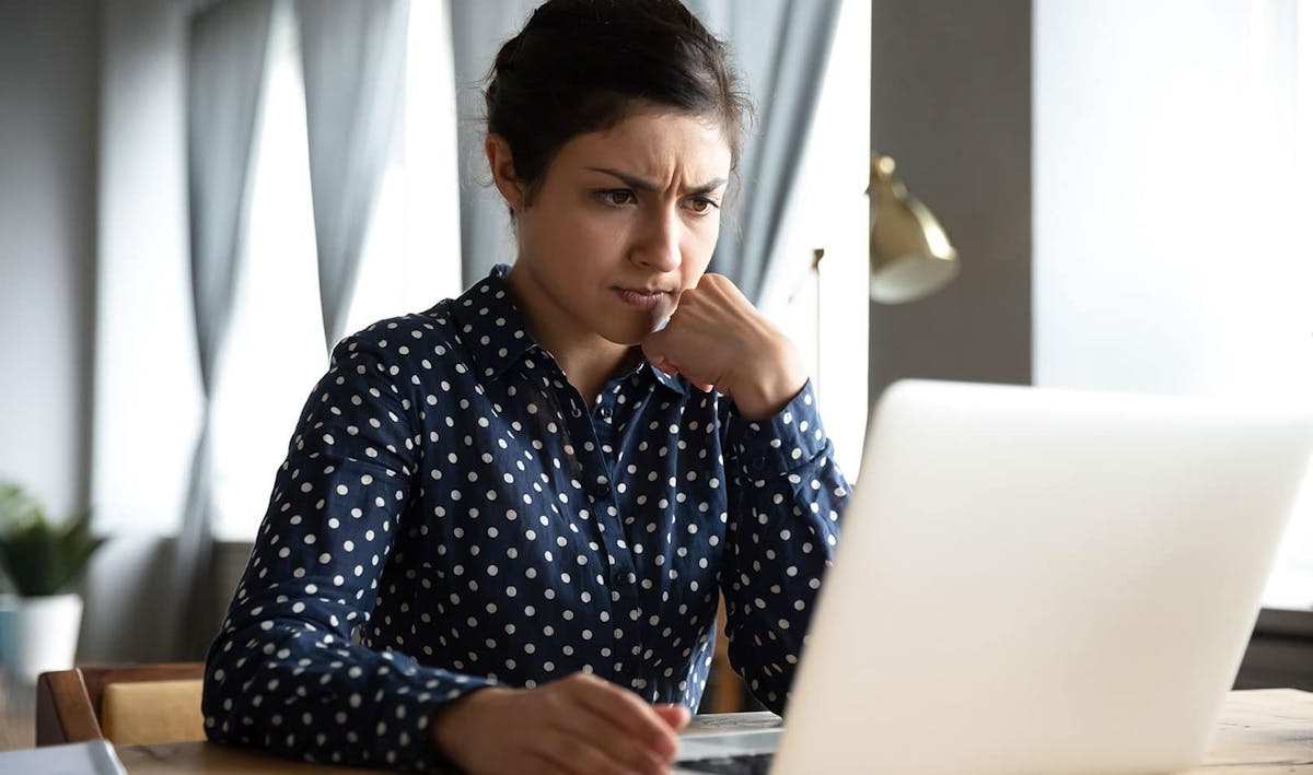 Ung dame med mørkt hår i hestehale sitter på et kontor ser bekymret på laptopen sin