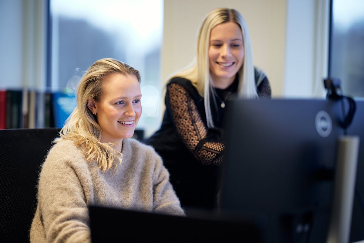 To smilende damer med lyst langt hår ser smilende på en pc skjerm der den ene tydelig hjelper den andre