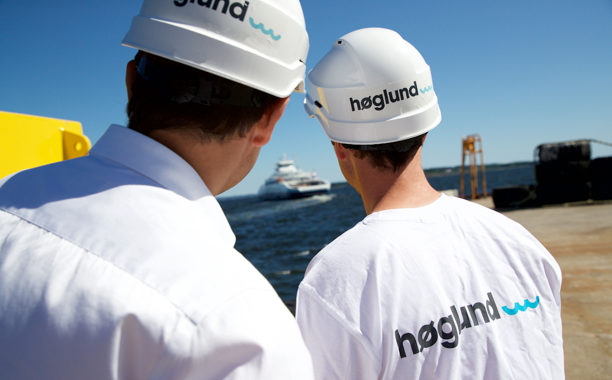 2 ansatte i Høglund står på en brygge med ryggen til kamera og ser ut på et skip som kommer mot dem. De har hvite hjelmer og t-skjorter med Høglund logo på