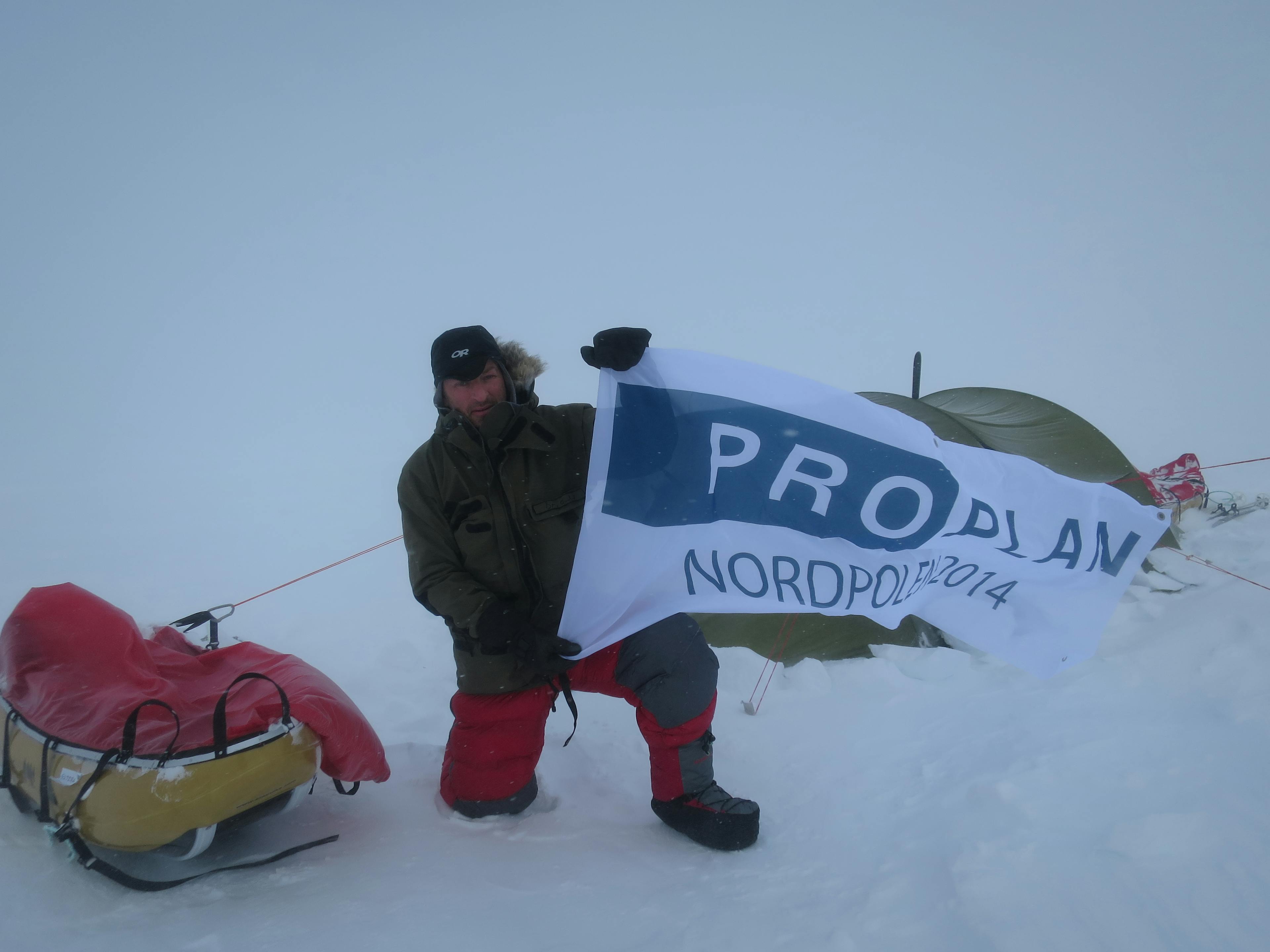 Bilde av en vinterkledd mann som holder et banner det står Proplan Nordpolen 2014 på
