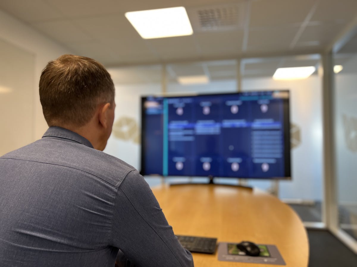 En mann i skjorte sitter ved et møtebord med ryggen til kamera og kikker på en stor skjerm med masse data