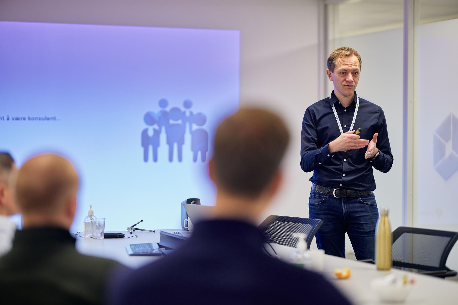 Mann står foran en presentasjonsskjerm og snakker til et publikum som vi bare ser konturene av