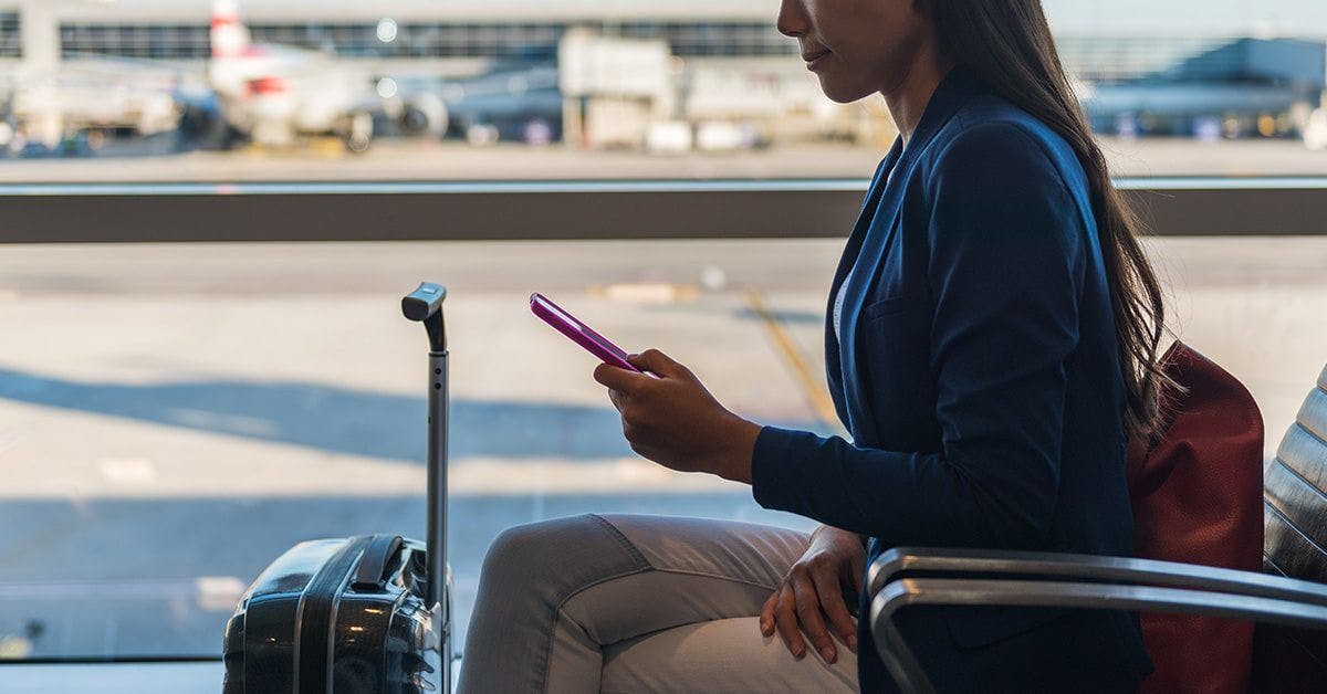 Dame som sitter på flyplass med en koffert fremfor seg og ser på mobilen der hun enkelt fører reiseregningen digitalt