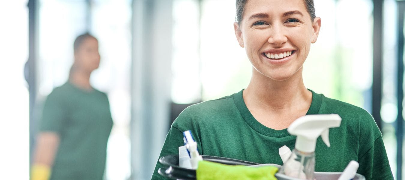 Smilende dame med brunt hår i hestehale og grønn t-skjorte holder en bøtte med rengjøringsmidler