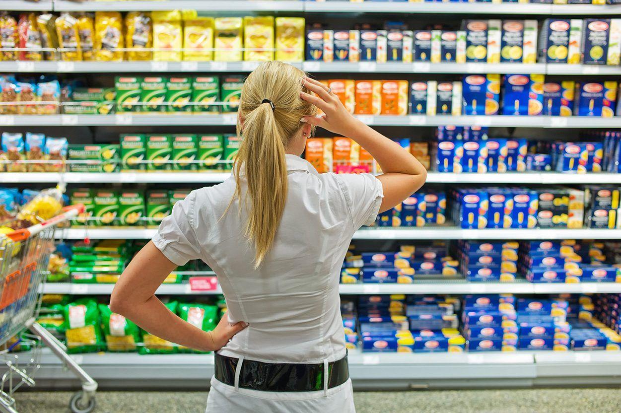 Ung dame med langt lyst hår i hestehale står i butikken foran pastahyllen og tar seg til hodet for hun aner ikke hva hun skal velge