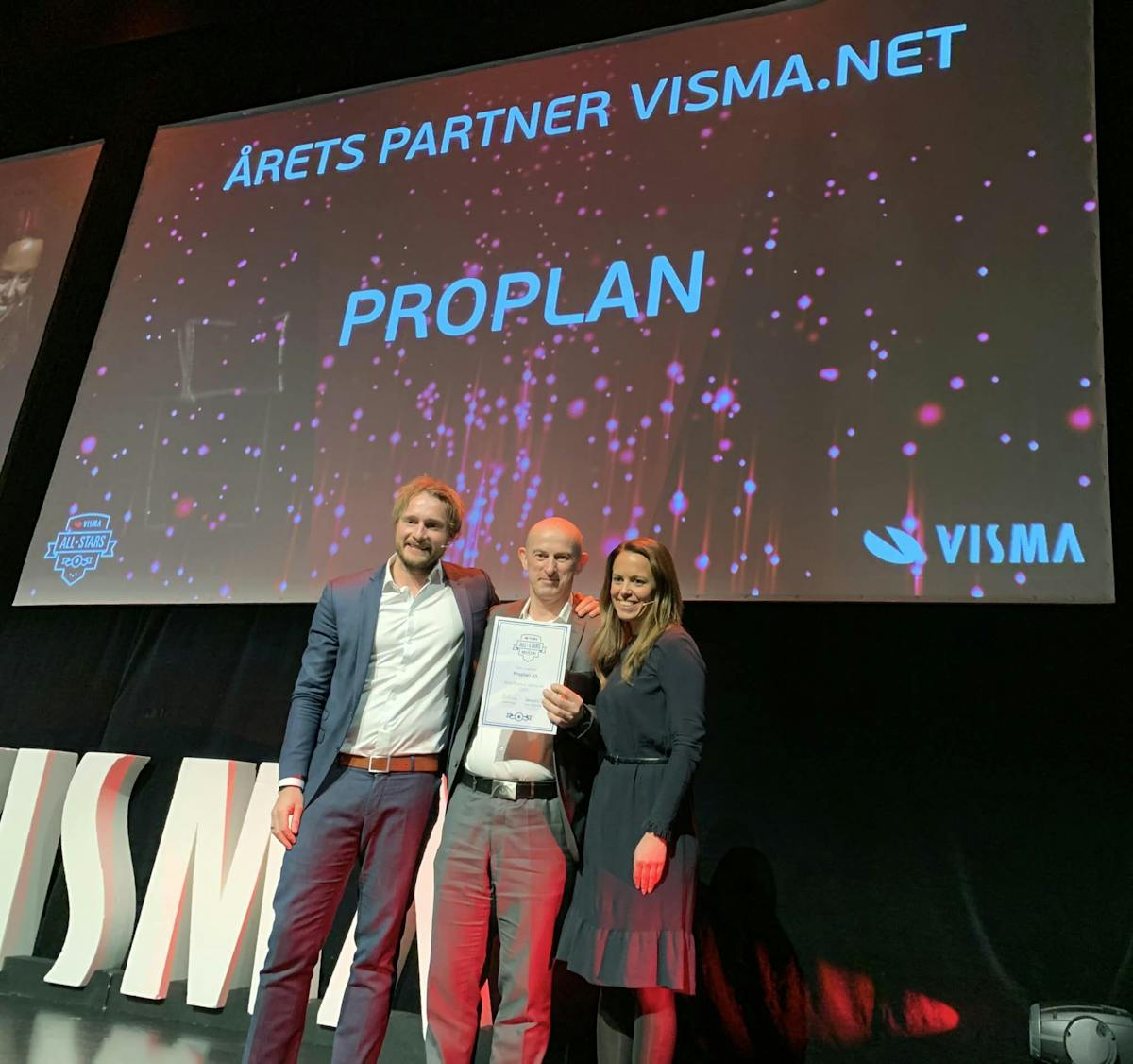To menn og en dame står på en scene der den ene mannen har mottatt en pris med en storskjerm i bakgrunnen der det står at Proplan er vinner av prisen årets partner visma.net